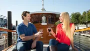 A man and a woman are smiling and toasting with glasses of red wine on a boat deck under clear skies.