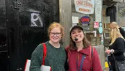 Two people are smiling in front of Yonah Schimmel's Knishery, which has various signs indicating social distancing rules and business hours.