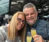 A smiling couple is posing for a photo while holding glasses of a yellow beverage in a dining area with a modern glass enclosure