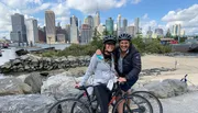 Two people wearing helmets are posing with a bicycle in front of a city skyline.