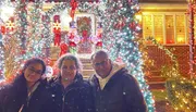 Three people pose for a photo in front of a house adorned with vibrant Christmas decorations and lights.