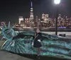 A person is posing next to a sculpture of a giant head resembling the Statue of Liberty with the illuminated New York City skyline in the background at night