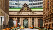 The image shows the historical facade of Grand Central Terminal in New York City, framed by modern buildings, with yellow taxis in the foreground.