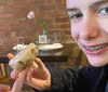 A smiling young person with braces holds up a cookie resembling a fortune cookie at a table with a brick wall backdrop