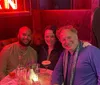 Three people are smiling for a photo while sitting in a booth with a neon sign in the background suggesting they are in a bar or restaurant