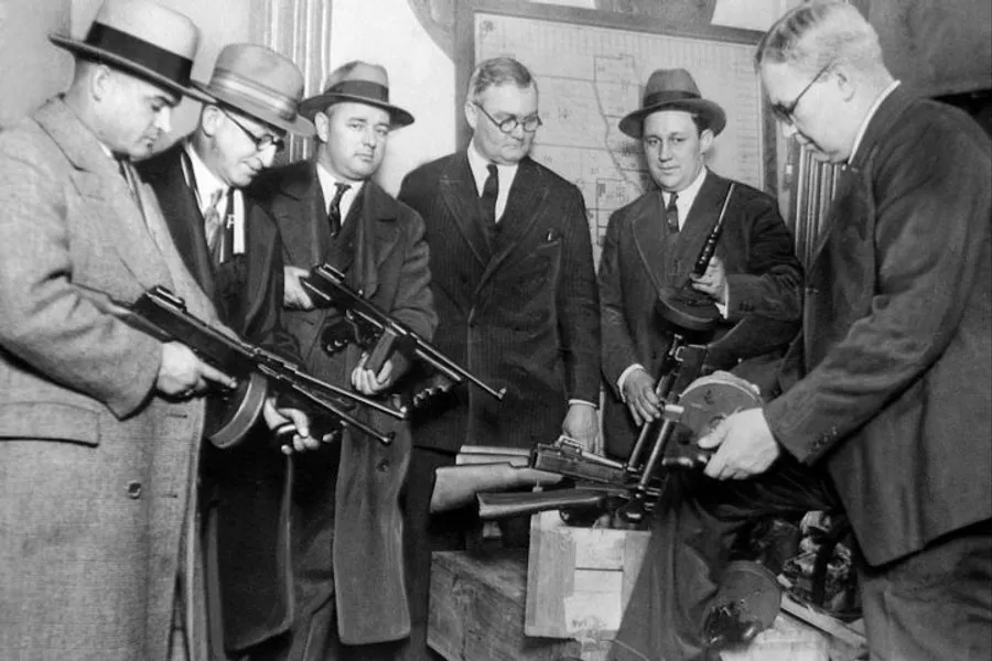 A group of men in suits and hats from an earlier era are examining or displaying a collection of Thompson submachine guns.