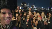 A group of smiling people are posing for a selfie with a nighttime cityscape in the background.