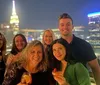 A group of people are posing for a photo with drinks at a rooftop venue at night with the illuminated skyline of a city in the background