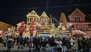 A bustling holiday scene where a crowd gathers in front of festively decorated homes illuminated by Christmas lights and flanked by giant nutcracker statues.