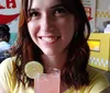 A smiling woman is holding a glass with a pink beverage garnished with a lime slice in a casual dining setting with vibrant decor