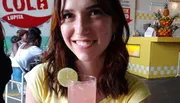 A smiling woman is holding a glass with a pink beverage garnished with a lime slice, in a casual dining setting with vibrant decor.