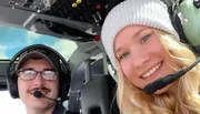 A man and a woman in aviation headsets are taking a selfie inside the cockpit of an aircraft, both smiling at the camera.