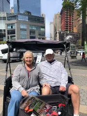 A smiling couple is sitting in a pedicab in an urban setting featuring modern buildings and a globe sculpture in the background.