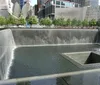 The image shows visitors at a reflective memorial with a weathered metal beam in the foreground and a cityscape in the background