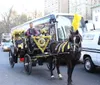 A horse-drawn carriage adorned with yellow and pink plumes and decorations is trotting through an urban street with passengers and a driver onboard amidst modern vehicular traffic