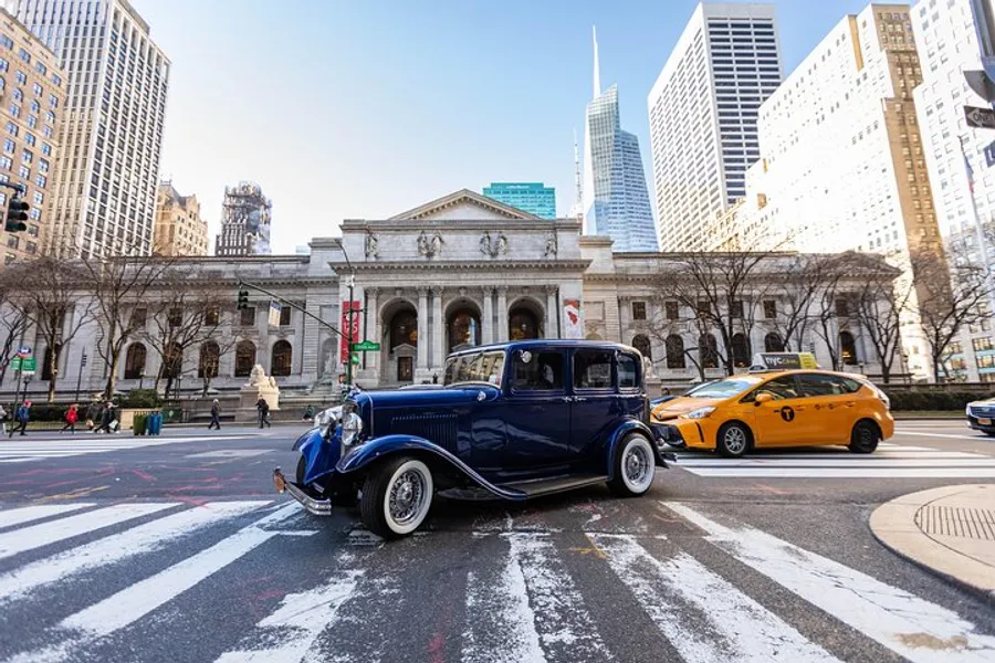 A vintage blue car is parked on a city street with pedestrian crosswalks, contrasting with a modern yellow taxi, in front of a stately building with classical architecture.