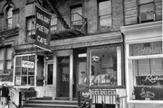 The image is a black and white photo of the exterior of the Gaslight Poetry Cafe, complete with signs and a person standing by the entrance.