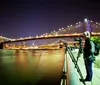 A person stands beside a camera on a tripod capturing a long-exposure photograph of a brightly lit suspension bridge at night with a city skyline in the background