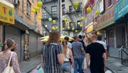 Pedestrians walk down a vibrant street adorned with yellow and green lanterns, showcasing the cultural atmosphere of a busy Chinatown district.