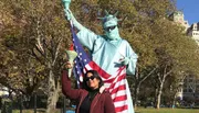 A person is posing with a person dressed as the Statue of Liberty, who is holding an American flag, in a park setting with trees in the background.