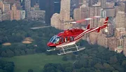 A red and white helicopter is flying over Central Park with the New York City skyline in the background.