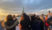 A group of people are capturing the view of the Statue of Liberty with their phones at sunset.