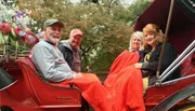 Four people are smiling for the camera while sitting in a red horse-drawn carriage, covered with a red blanket, and adorned with colorful artificial flowers.