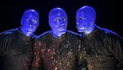 Three performers with blue-painted faces and speckled black shirts appear illuminated under a dark background.