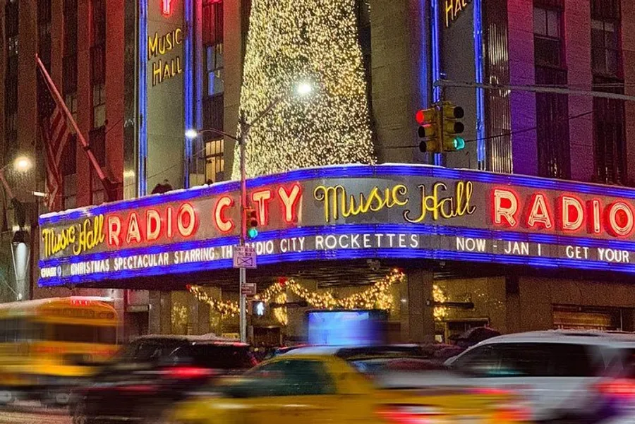 The image displays the vibrant, illuminated marquee of Radio City Music Hall at night with blurred traffic in the foreground, suggesting the bustling energy of an urban environment.
