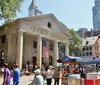 People are gathered around Quincy Market on a sunny day with street vendors and an American flag adorning the facade