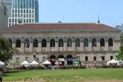 The image shows a stately building with arched windows and flags, set against a clear sky with some trees and tents in front, suggesting a public or historical venue, possibly a library or government edifice.