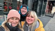 Three people are smiling for a selfie on a city street, with decorations in the window behind them suggesting it might be around Valentine's Day.