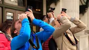 A group of people are looking up, each holding binoculars or cameras to their eyes, seemingly focused on something out of the frame.