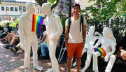 A smiling person stands beside white statues of people, one of which is holding a rainbow pride flag, in what appears to be a vibrant outdoor setting.