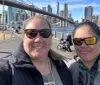 Two people are taking a selfie with the Brooklyn Bridge and the New York City skyline in the background