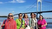 A group of smiling women poses in front of a suspension bridge on a sunny day.