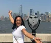 A person is posing with their arm extended simulating touching the top of the One World Trade Center across the river with the New York City skyline in the background