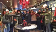 A group of smiling people are holding hands in a line while posing for a photo inside a cozy shop decorated with winter-themed items and a chandelier.