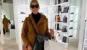 A person is smiling for the camera inside a boutique with bags and shoes on display, wearing sunglasses and a teddy coat, and holding a shopping bag.