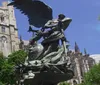 The image shows a dramatic bronze statue with an angelic figure in front of an ornate Gothic-style cathedral and lush green trees under a clear blue sky