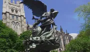 The image shows a dramatic bronze statue with an angelic figure in front of an ornate Gothic-style cathedral and lush green trees under a clear blue sky.