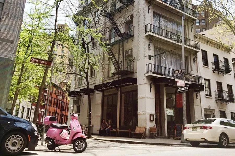 This image shows a sunny street corner with a pink scooter parked in front of a building with a cafe at street level, pedestrians sitting outside, and street signs indicating Christopher and Gay St.