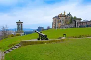 The image displays the historic Calton Hill in Edinburgh, Scotland, with its iconic monuments and an old cannon, under a cloudy sky.