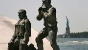 The image features bronze statues of military figures foregrounded against the backdrop of the Statue of Liberty in the distance.