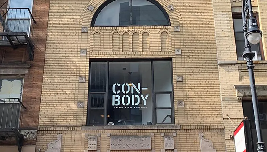 The image shows the facade of a building with a large window featuring the words CONBODY: PRISON STYLE BOOTCAMP, suggesting a workout facility offering a fitness program inspired by prison routines.