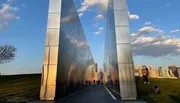 The image shows a modern, reflective monument pathway with people walking through it, overlooking a city skyline in the background during sunset.