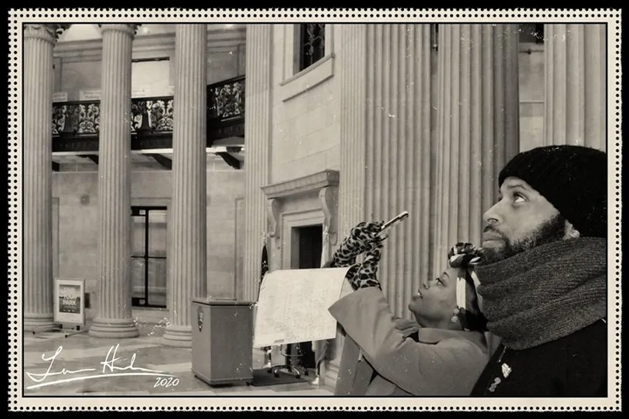 The image shows two people, one holding a paper looking up towards something unseen with a look of concentration, in a setting that resembles a museum or institution with classical architecture, all presented in a sepia tone possibly to evoke a bygone era.
