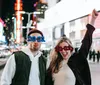 A man and a woman are posing comically in a brightly lit urban setting at night wearing novelty glasses that say 2021