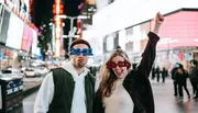 A man and a woman are posing comically in a brightly lit urban setting at night, wearing novelty glasses that say 