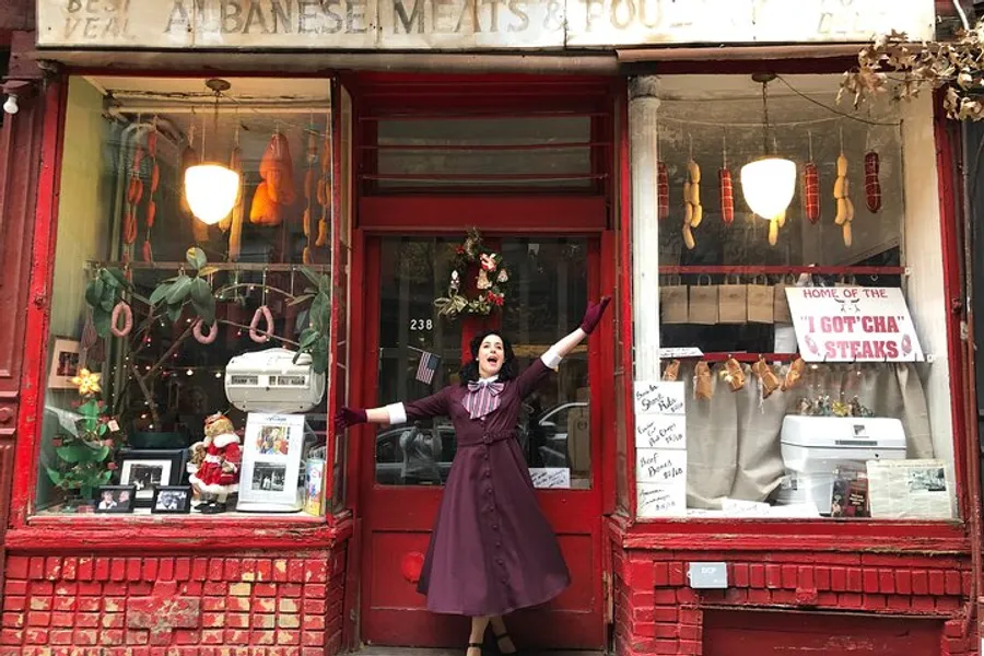 A person in a clown-like outfit is cheerfully posing with arms outstretched in front of an old-fashioned red storefront adorned with holiday decorations and a sign for Albanese Meats & Poultry.